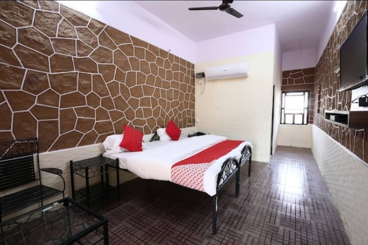 A bedroom with a stone-patterned wall, a bed with red pillows, a ceiling fan, and a TV mounted on the wall.