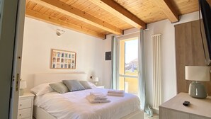 Una camera da letto con soffitto in legno, un letto con biancheria bianca, un comodino, una lampada e vista sull'esterno.