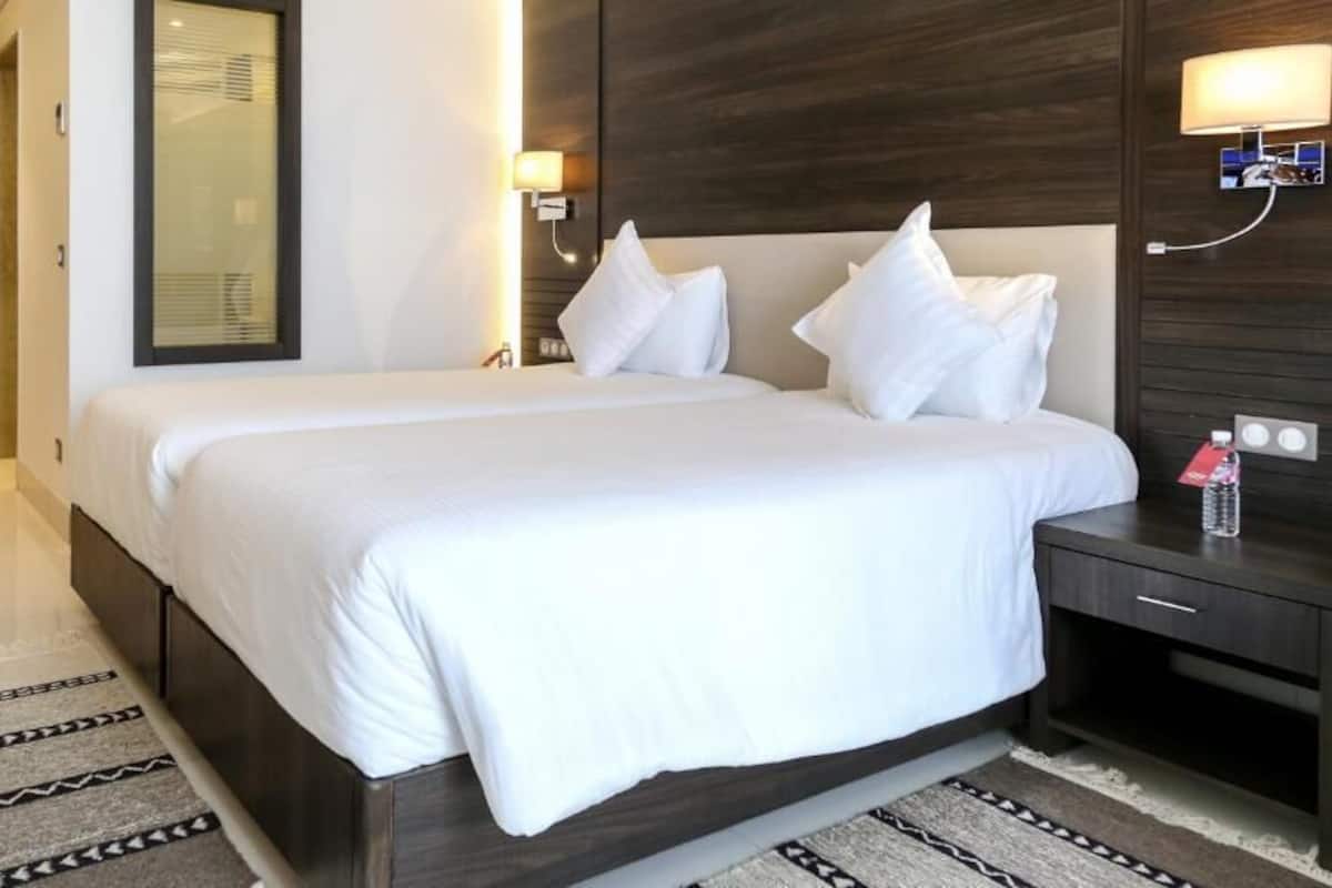 A hotel room with a large bed, a bedside table, a nightstand, a wall-mounted lamp, and a door.