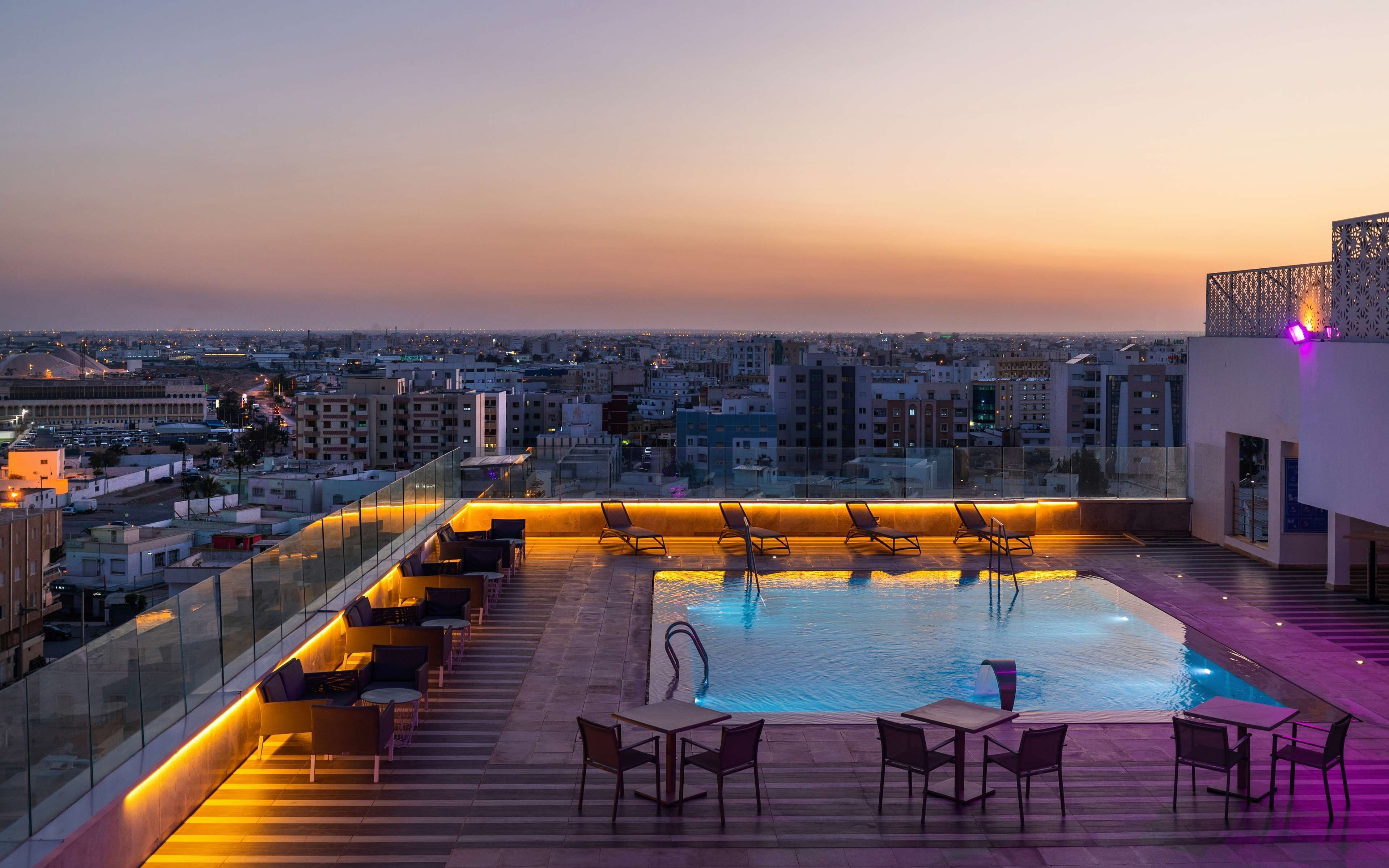 Un área de piscina en la azotea con asientos de descanso y vistas a la ciudad al anochecer.