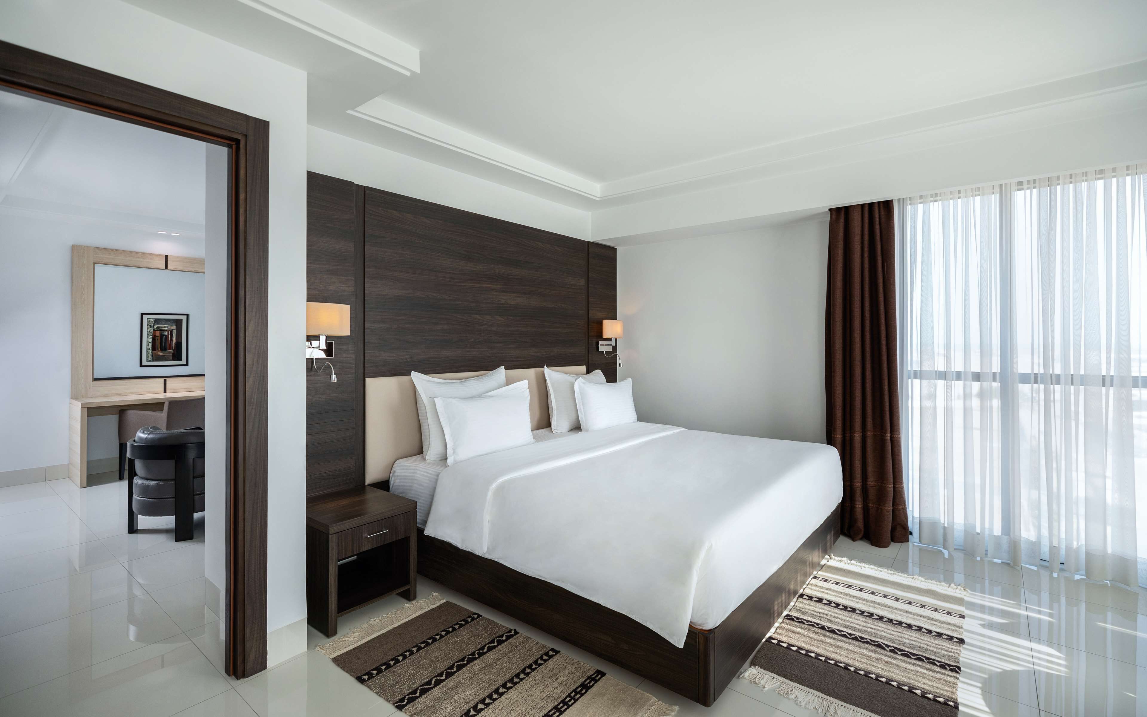 A modern hotel room with a large bed, dark wood headboard, two bedside lamps, a desk with a chair, and a large window with curtains.