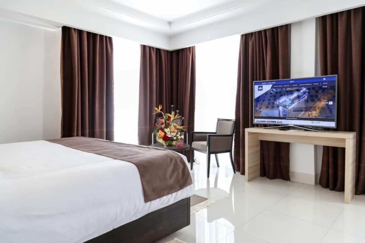 A hotel room with a bed, a desk with a television, and a vase of flowers.