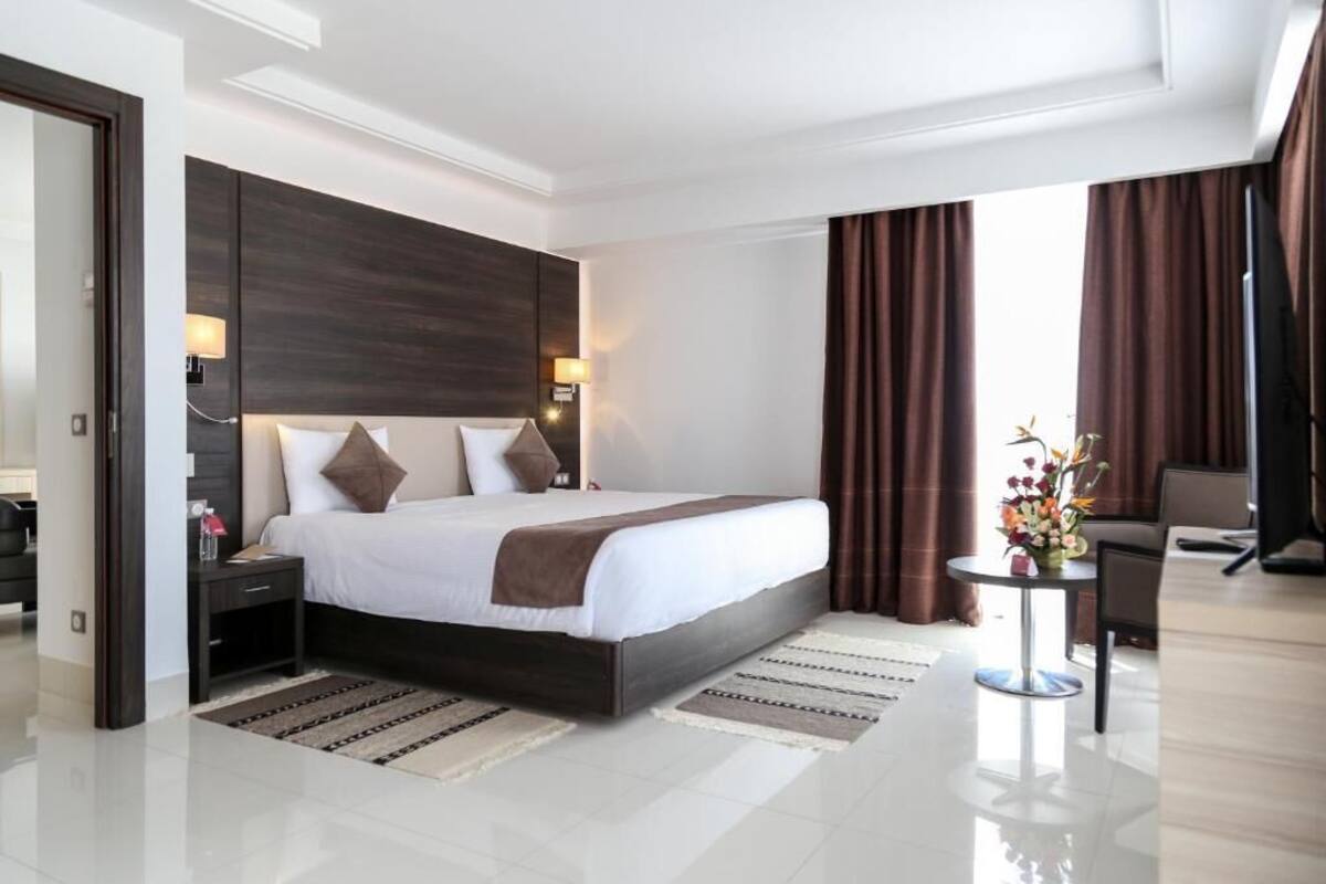 A modern hotel room with a large bed, dark wood headboard, bedside lamps, a small table with flowers, and a view of the outdoors through curtains.