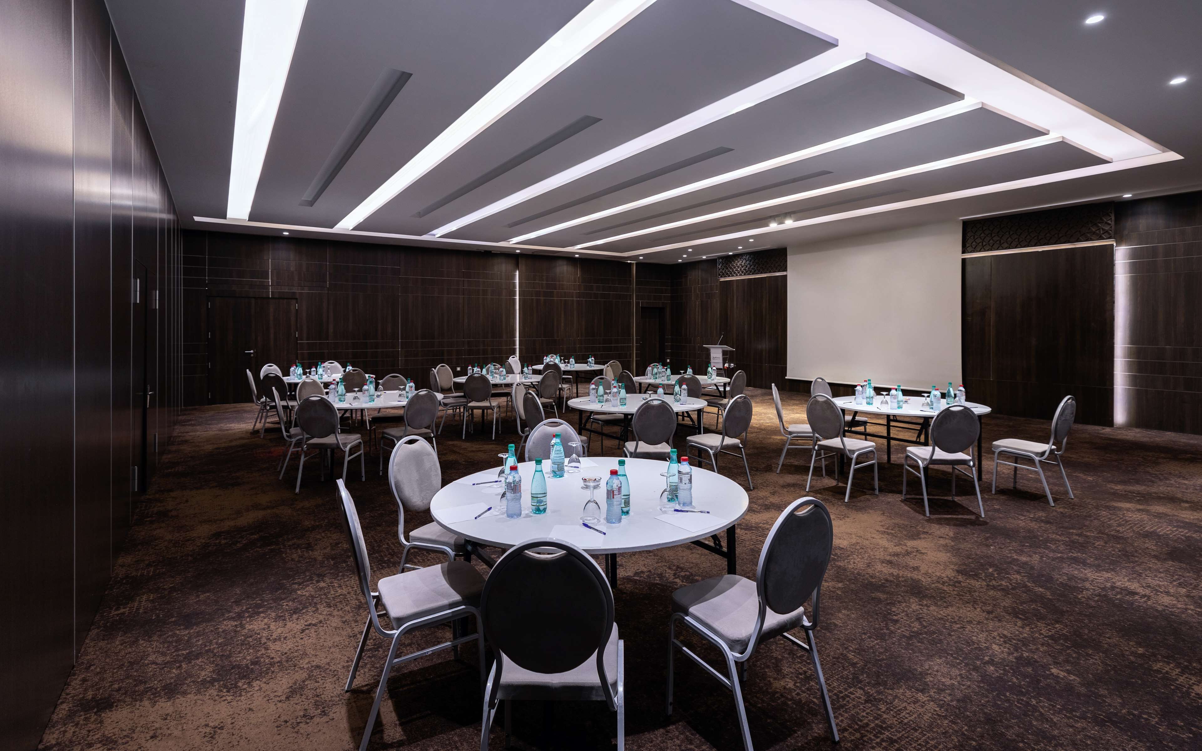 Un salón de conferencias con mesas redondas, sillas y botellas de agua, con un diseño de techo moderno y paneles de madera oscura.