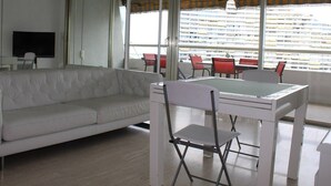 A modern living room with a white sofa, a dining table with chairs, and a large window with blinds.