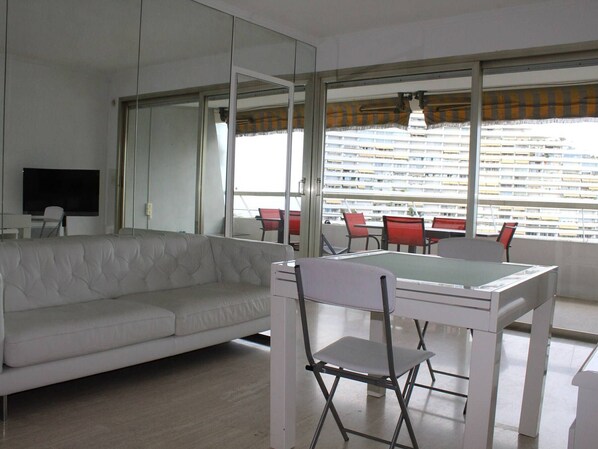 A modern living room with a white sofa, a dining table with chairs, and a large window with blinds. - Villeneuve-Loubet: Apartment with Terrace, Air Conditioning and Garage (Villeneuve-Loubet)