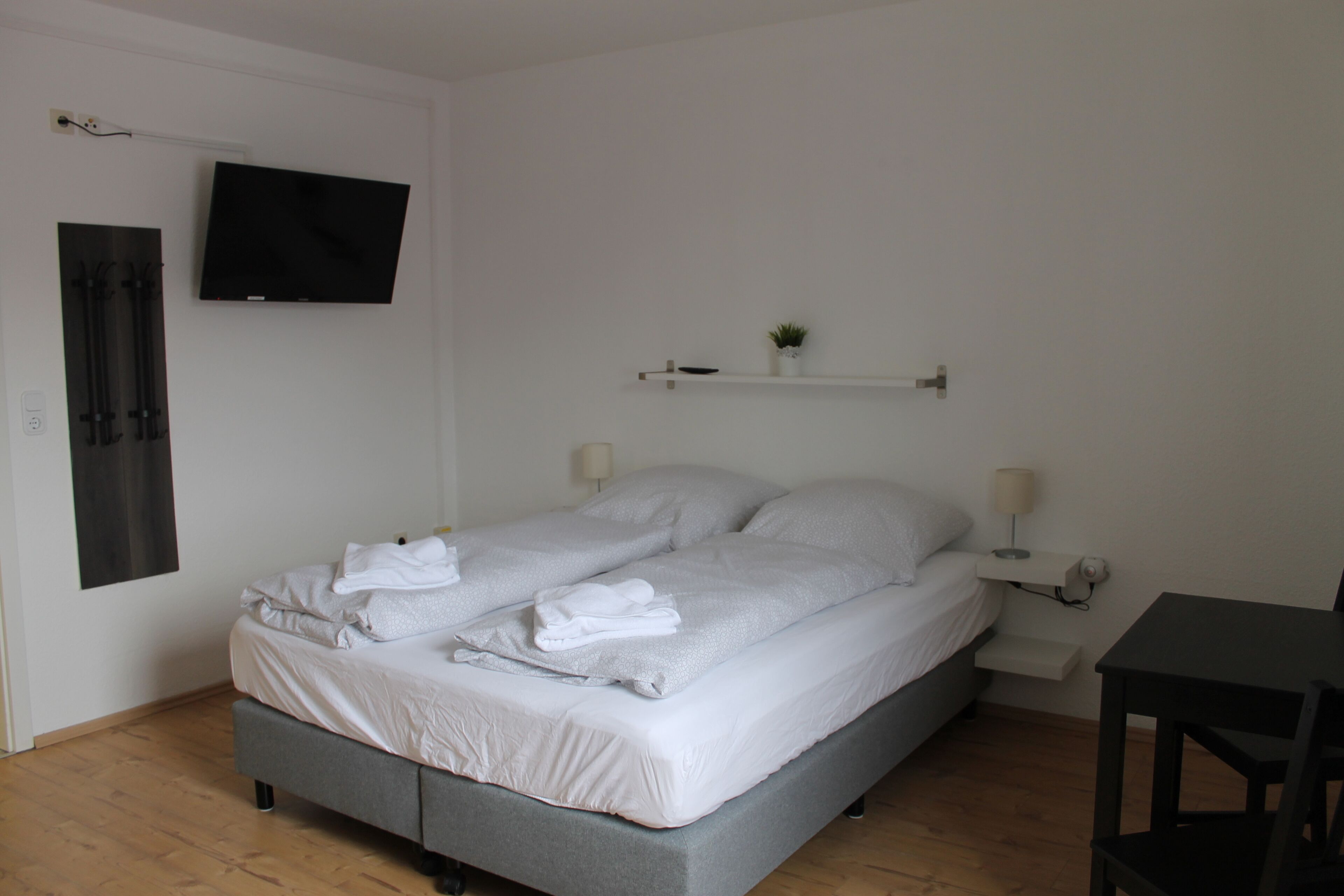 A neatly made bed with white linens and towels, a flat-screen TV mounted on the wall, a small shelf with a plant, and a desk with a chair.