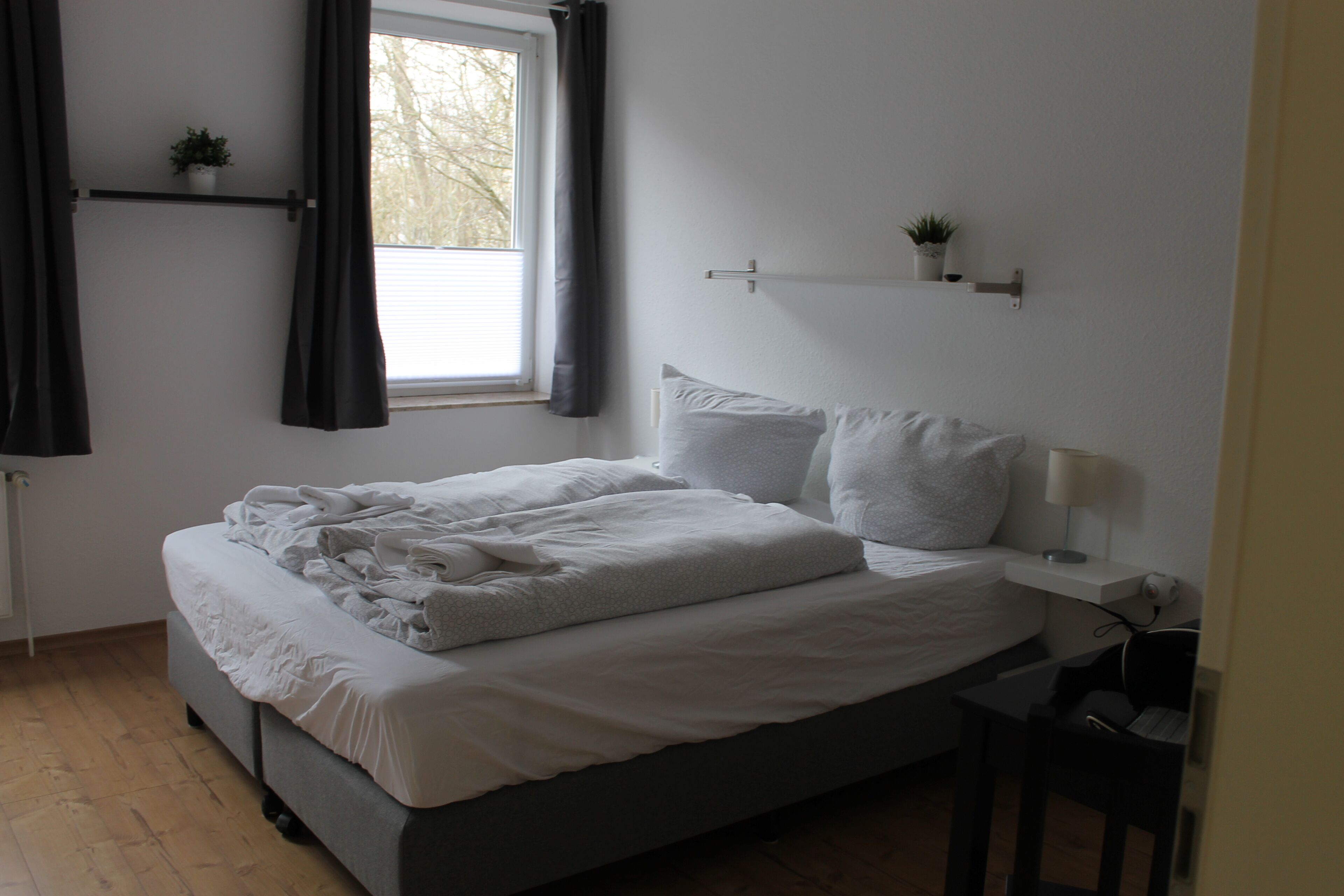 Una cama bien hecha con sábanas y almohadillas blancas, una mesita de noche con lámpara y una ventana con cortinas.