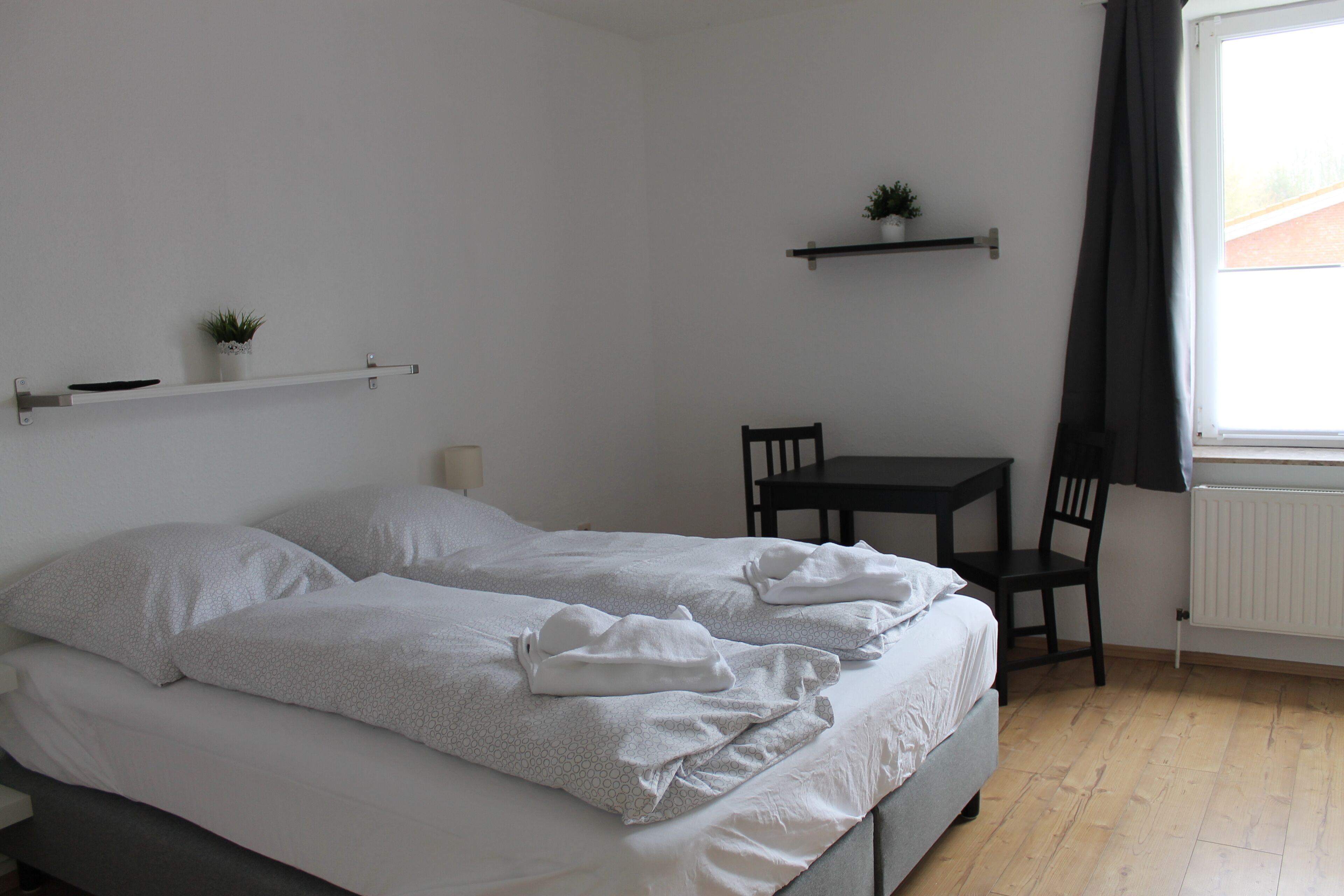 Un dormitorio con cama, mesita de noche, silla y ventana con cortinas.