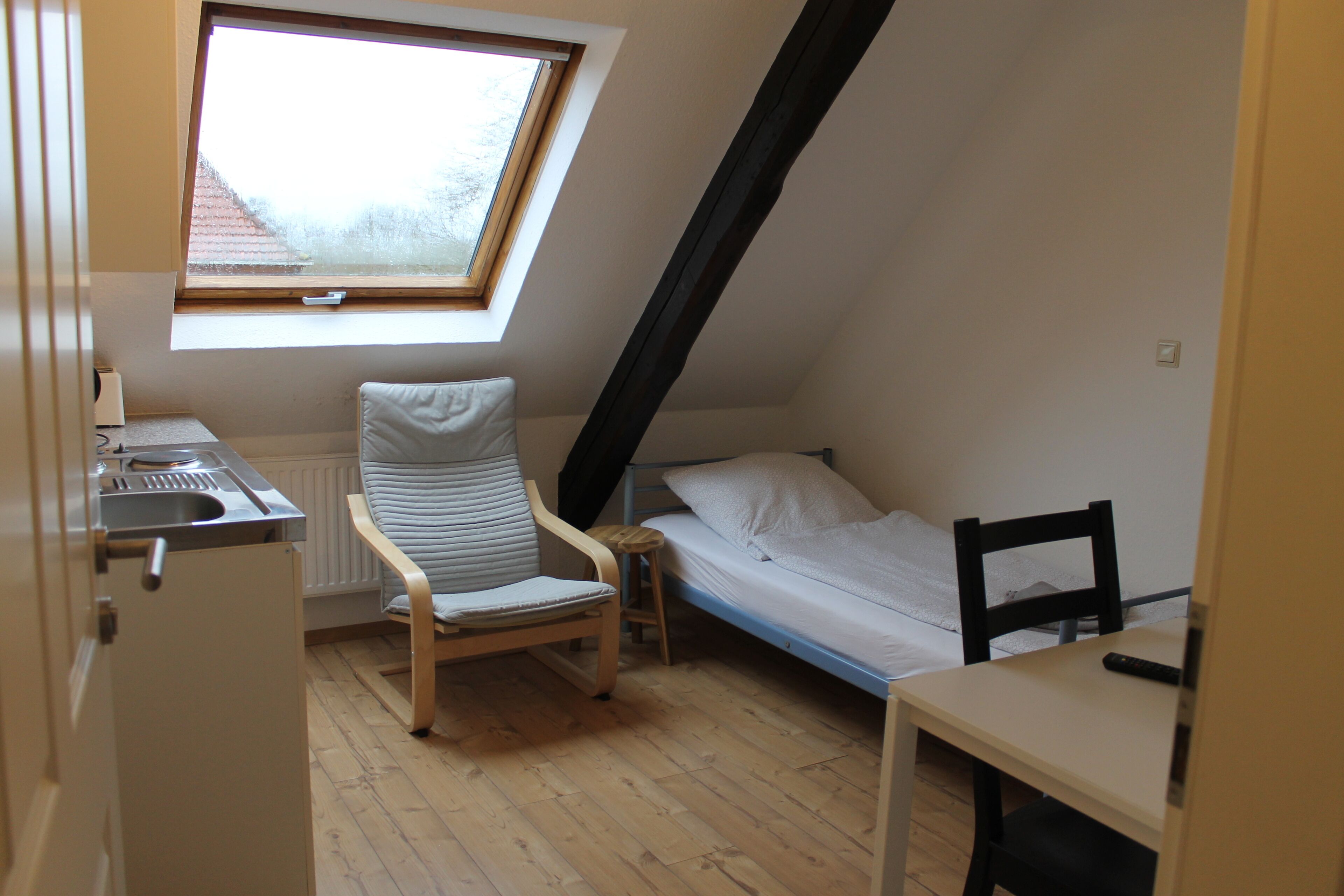 Una habitación pequeña y acogedora con techo inclinado, una cama individual, una silla, una mesita y una ventana con vistas a un edificio.