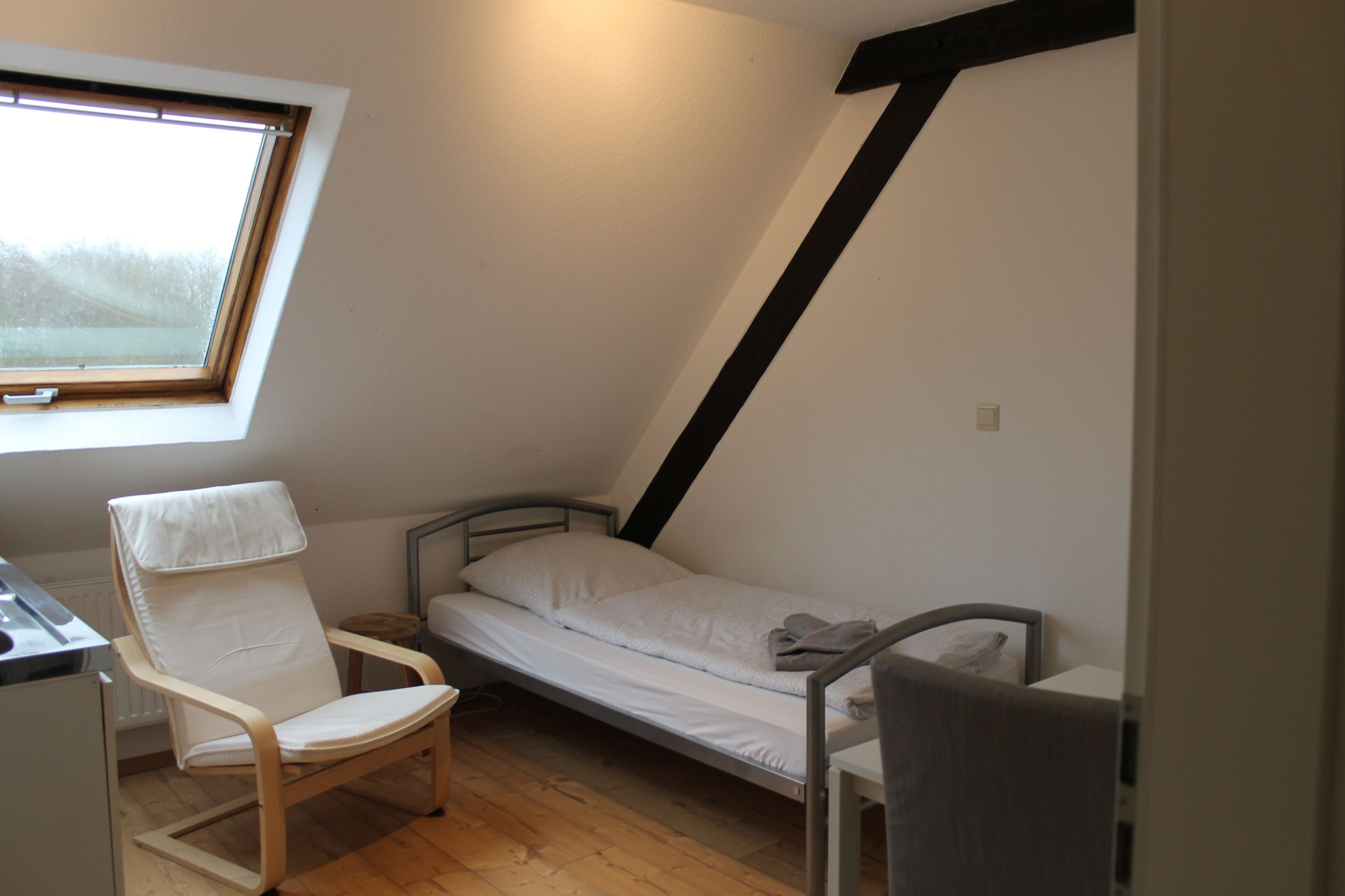 Een kleine kamer met een enkel bed, schuine wand, raam, stoel en bureau.