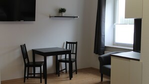 A small room with a wooden floor, a table with two chairs, a TV mounted on the wall, a shelf with a plant, and a window with curtains.