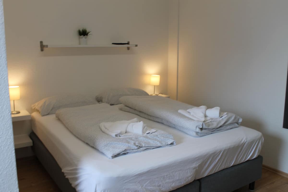 Uma cama bem arrumada com lençóis brancos e um cobertor cinza, duas luminárias de cabeceira e um pequeno prateleira com uma planta.