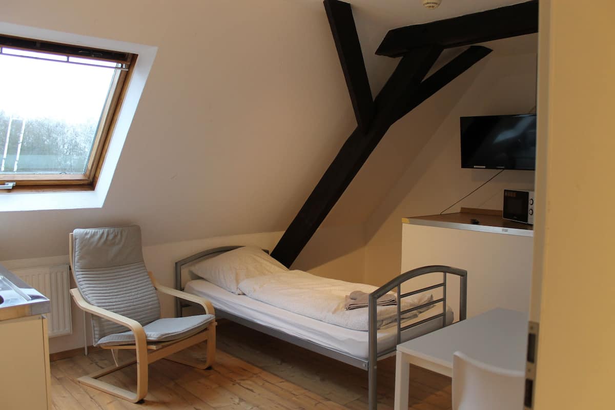 Quarto pequeno com uma cama de solteiro, teto inclinado, escrivaninha, cadeira e televisão.