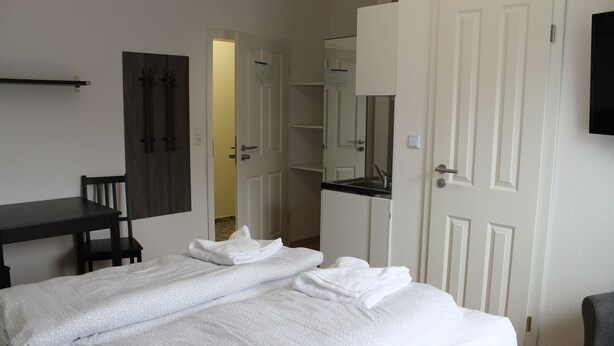 A neatly made bed with white linens, a small desk with a chair, a dark wooden wardrobe, and a white door with a mirror above it.