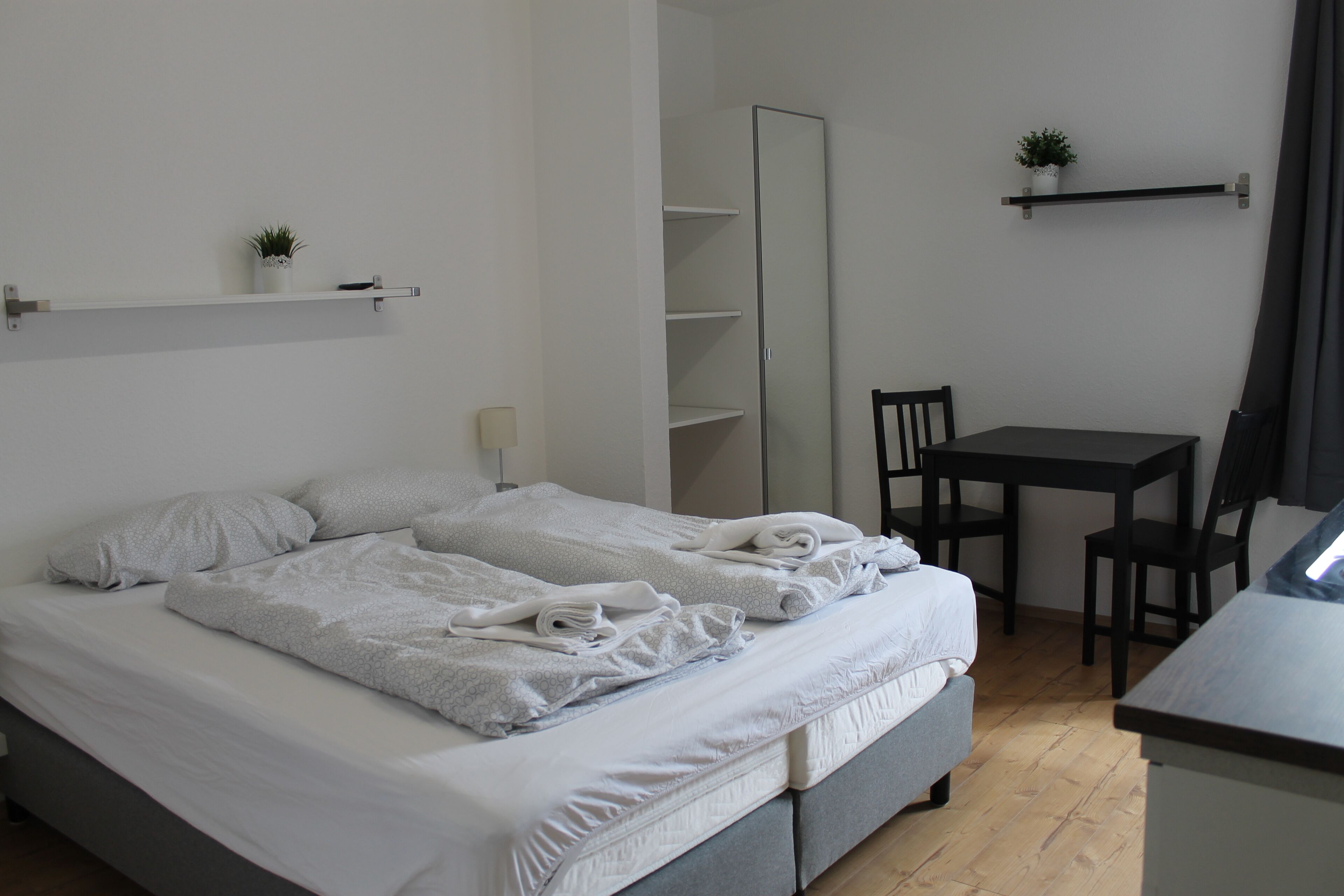 A neatly made bed with white linens and towels, a small bedside table, a chair, a desk, and a mirror.