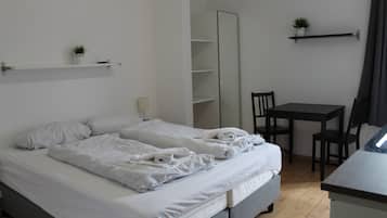 Una cama bien hecha con sábanas y toallas blancas, una mesita de noche pequeña, una silla, un escritorio y un espejo.