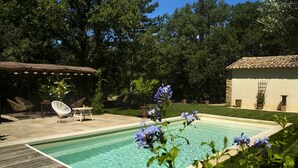 A swimming pool with a wooden deck, surrounded by lush greenery and a small patio area with outdoor furniture. - L'Atelier, with its century-old oak trees and panoramic view of the Luberon region (Cucuron)