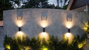 A modern building facade with three illuminated wall-mounted lights and a landscaped garden area with shrubs. - The Elegance Pool Villas at KP Beach (Ko Chang)