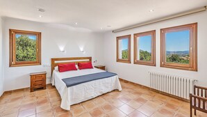 Une chambre à coucher comprenant un lit, deux tables de chevet, une chaise et trois fenêtres donnant sur un paysage agréable.