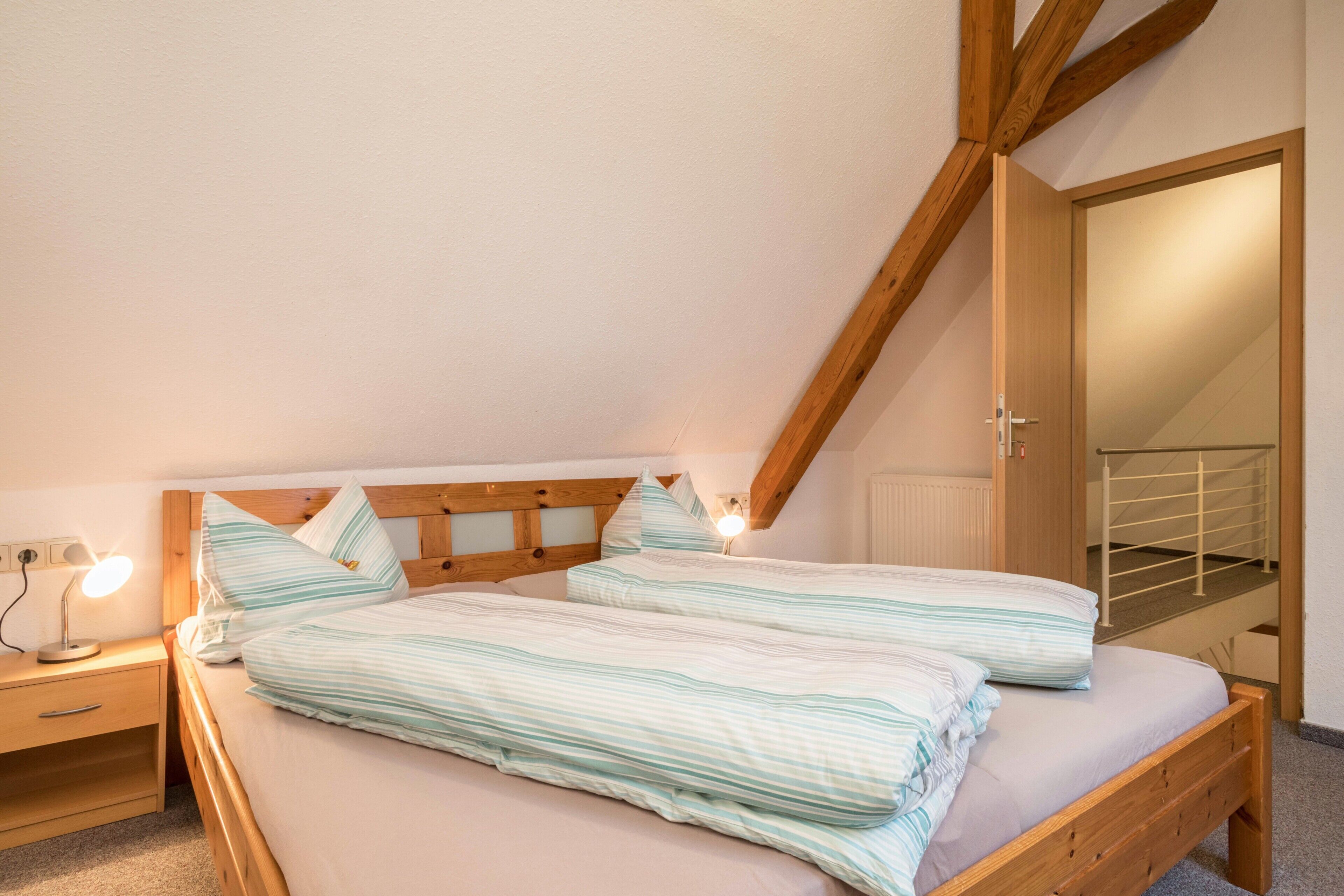 Une chambre avec une tête de lit en bois, deux lits avec une literie à rayures, une table de chevet avec une lampe, et une vue sur un escalier menant à une porte.