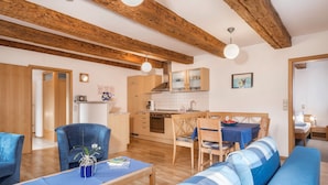 A cozy living room with a sofa, a dining table, and a kitchen area with wooden beams and cabinets. - Apartment “4-Star Holiday Apartment No. 3” with Wi-Fi, Balcony, Garden & Terrace (Deggenhausertal)