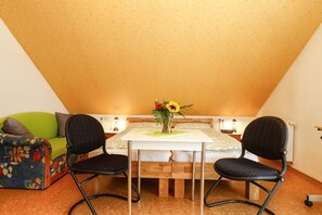 A small, cozy room with a sloped ceiling, a green sofa, a wooden table with two chairs, and a vase with flowers.