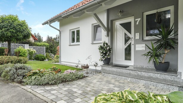 Un bâtiment à la façade blanche, doté d’un toit en tuiles et portant le chiffre « 7 » sur la porte. On y trouve des plantes en pot et un jardin bien entretenu à l’avant.