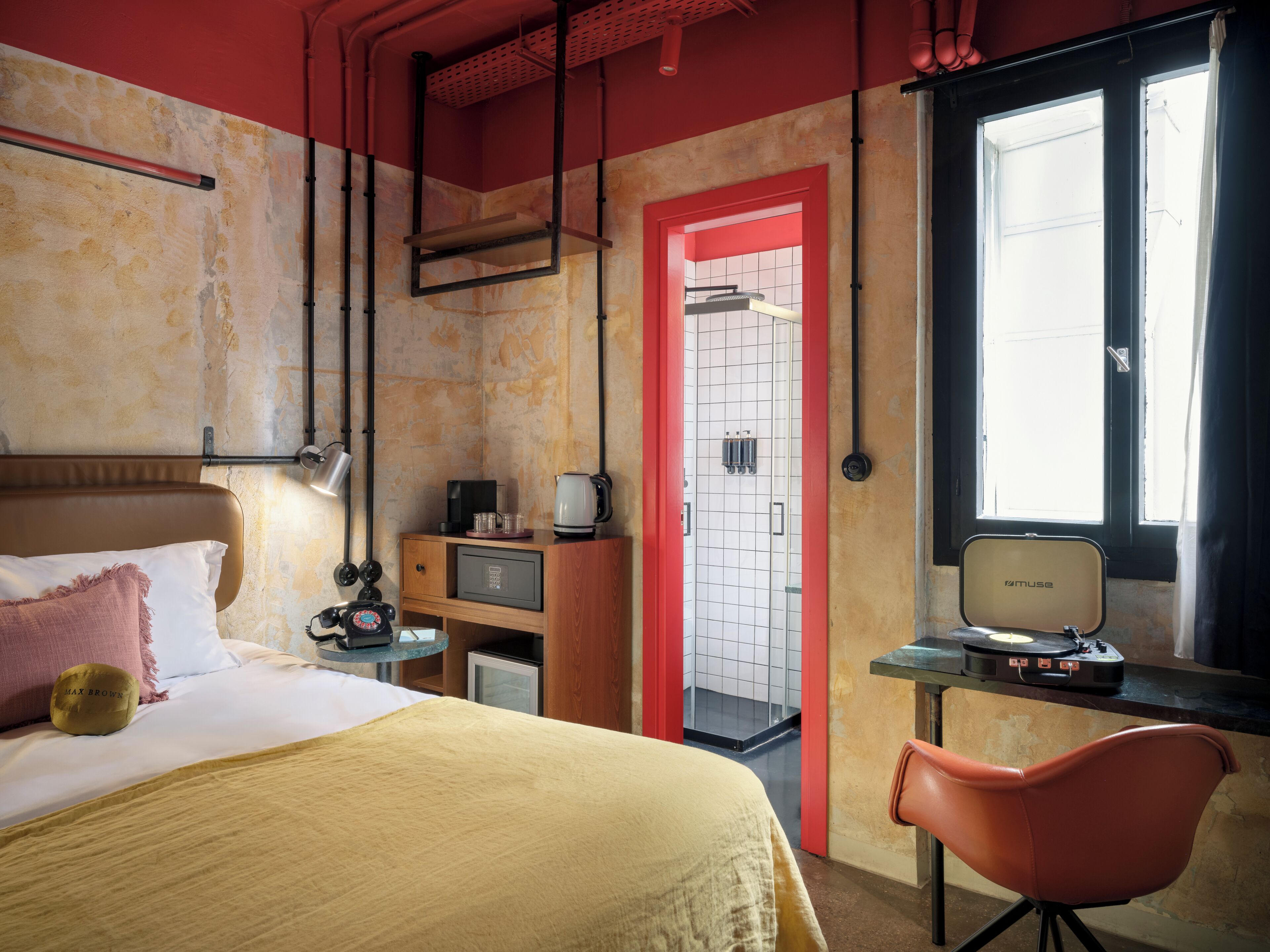 A hotel room with a bed, a desk with a computer, a chair, a red door, and a window.