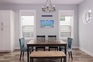 A dining area with a wooden table, four chairs, and a bench. There are two windows with blinds, a chandelier, and a painting of a beach scene. - Free Golf Cart, Walk to Beach, Pool Access, Sleeps 10, Bike to Shops & Restaurants! (Santa Rosa Beach)