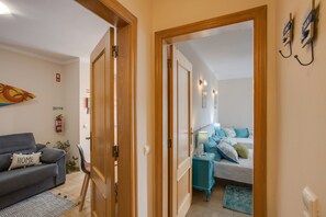 A room with a bed, a bedside table with a lamp, a sofa with a cushion, and a door leading to another room.