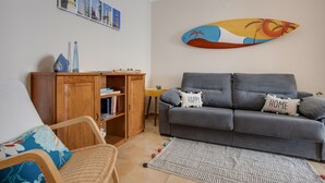 Ein Wohnzimmer mit grauem Sofa, Holzschrank, Korbstuhl und einem gemusterten Teppich.