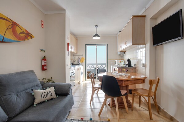 A living room with a sofa, a dining table with chairs, a kitchen area, and a large window.