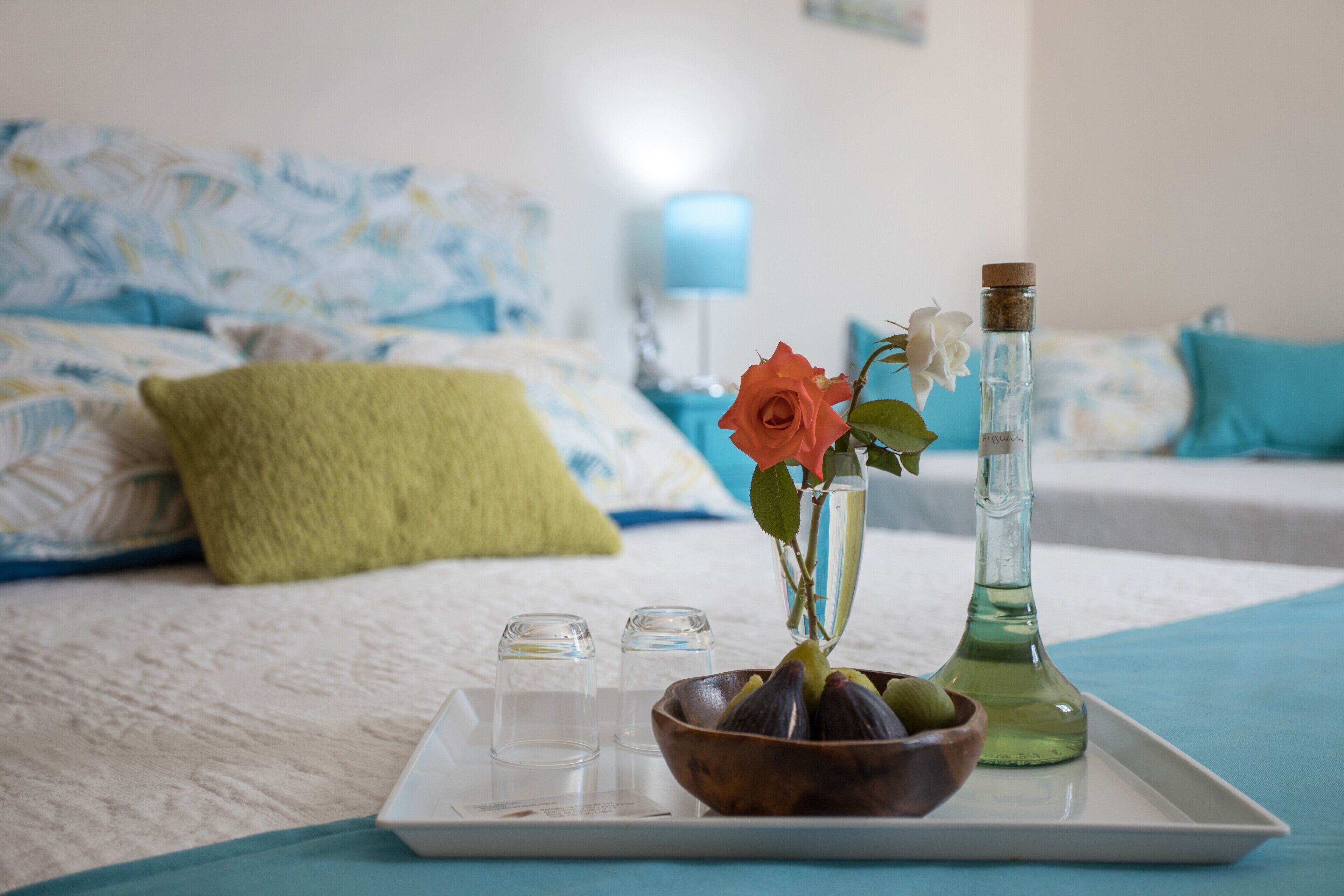 A tray with a bowl of figs, a rose, and a bottle on a bed with a blue blanket and pillows.