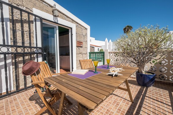 Une table et des chaises en bois sur une terrasse carrelée, avec vue sur un bâtiment et un arbre.