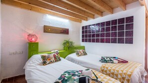 Una habitación con dos camas, un cabecero verde, una planta en la pared y un ventanal con cristales de color morado.