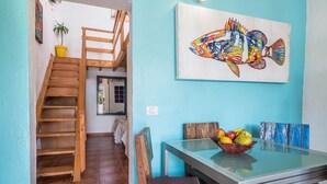 Un comedor con una mesa de vidrio, sillas de madera, una cesta de frutas y un cuadro de un pez colorido en la pared.