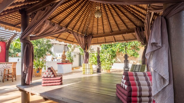 Un área de estar al aire libre con una mesa de madera, cojines y una estructura de techo de paja.