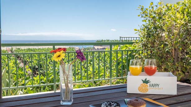 Un balcón con vistas a la playa, una mesa con una bandeja de pasteles, un jarrón con flores y una caja con la etiqueta 'HAPPY SUMMER'.