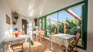 A bright room with wooden flooring, tables with floral tablecloths, and large windows with a view of greenery. - Cosy Holiday Home "Casa Sabina" in Alcalá with Terrace, Sea Access and Garden (Guía de Isora)