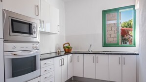 A kitchen with white cabinets, a microwave, an oven, a sink, and a window with a green frame. - Cosy Holiday Home "Casa Sabina" in Alcalá with Terrace, Sea Access and Garden (Guía de Isora)