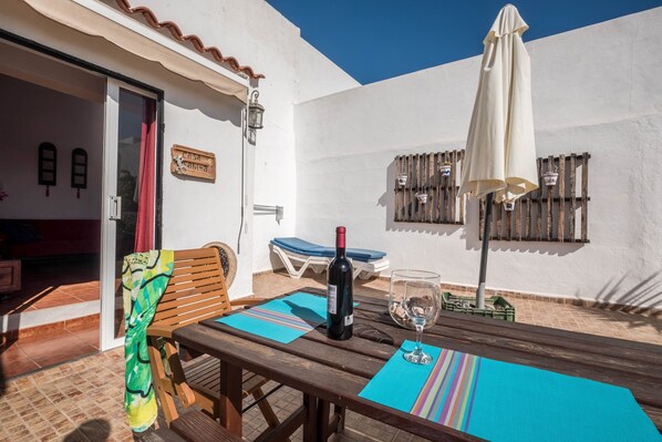 Un patio con una mesa de madera, una botella de vino y una copa. Hay un banco y una silla, y una puerta que da a un espacio interior.