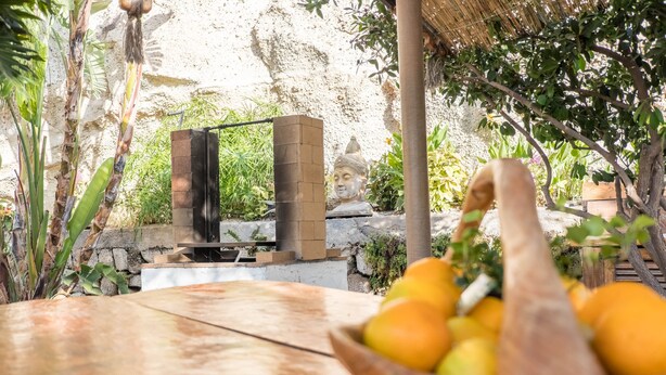 Une table en bois avec un panier de fruits, un mur de pierre et une statue de Bouddha en arrière-plan.