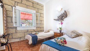 Une chambre avec deux lits, une fenêtre donnant sur un beau paysage, un mur en pierre, une sculpture décorative représentant un poisson et une petite table avec une plante.