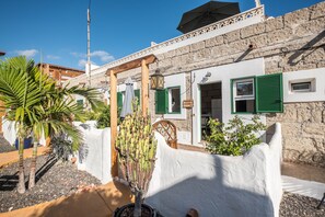 Un bâtiment blanc avec des volets verts, une porte en bois et un petit jardin agrémenté d’un cactus et d’un palmier.