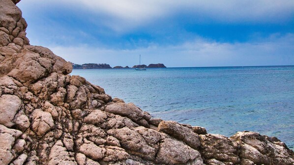 Une côte rocheuse bordée d’une eau d’un bleu limpide, avec un voilier visible au loin.