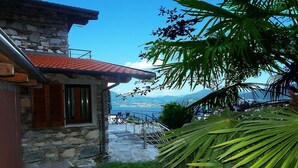 A stone building with a red-tiled roof, a staircase leading to a terrace, and a view of the sea and mountains.