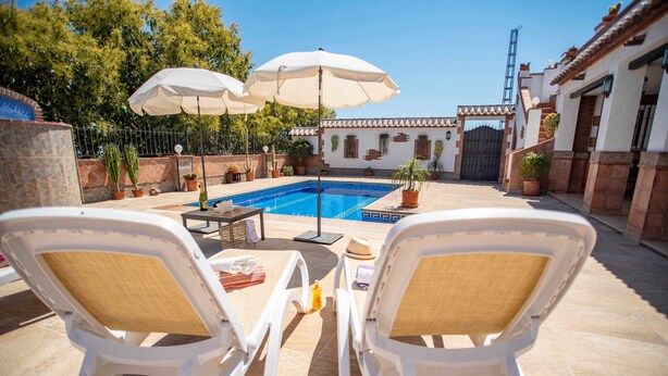 Uma área à beira da piscina com duas cadeiras de espreguiçadeira brancas, uma mesa e guarda-sóis.