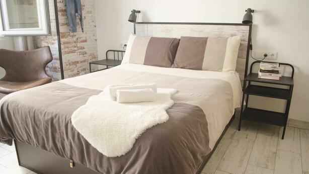 A neatly made bed with a fluffy white throw blanket and two pillows, a bedside table with a lamp, and a chair in a room with a brick wall and a window.