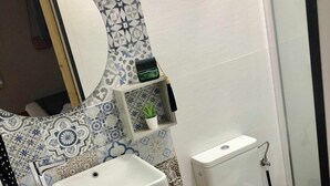 Ett badrum med ett vitt handfat, en toalett och en dusch. Ovanför handfatet finns en spegel, en liten hylla med en växt och en svart papperskorg.